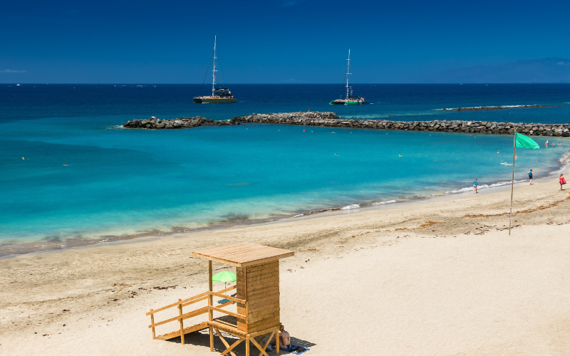 Une vue panoramique sur la plage de Playa del Duque, Tenerife, avec du sable doré et une cabane de sauveteur sur le côté gauche, affichant un drapeau vert. Concept : vacances à la plage, loisirs et tourisme à Tenerife